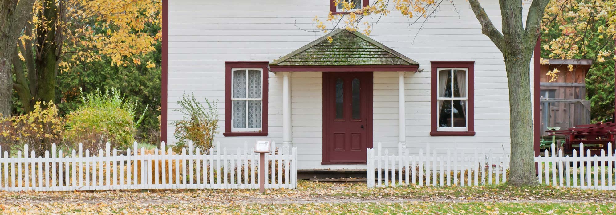 white picket fence in front of a white house with a red door and red window trim during fall